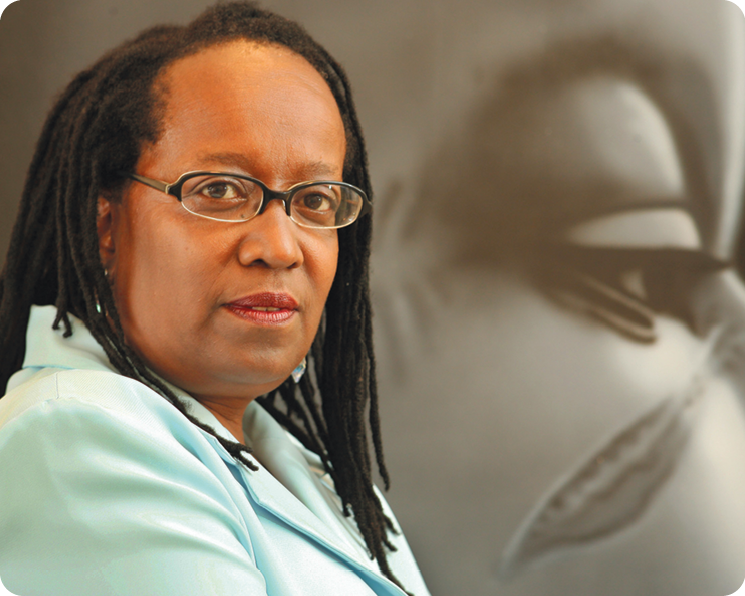 Fotografia. Destacando o busto de uma mulher negra usando óculos e terno. Ela está de perfil. Atrás, fotografia em preto e brancos destacando a face de uma pessoa negra.