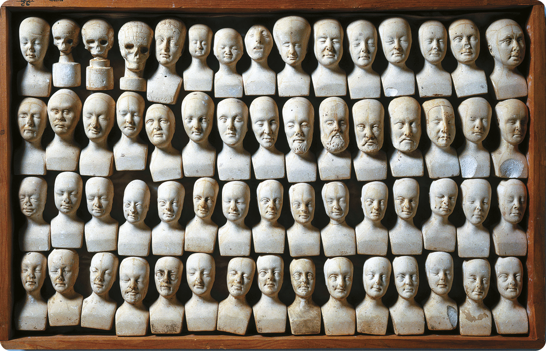 Fotografia. Diversas esculturas em miniatura de bustos de pessoas, uma do lado da outra. Elas estão empilhadas em quatro colunas.