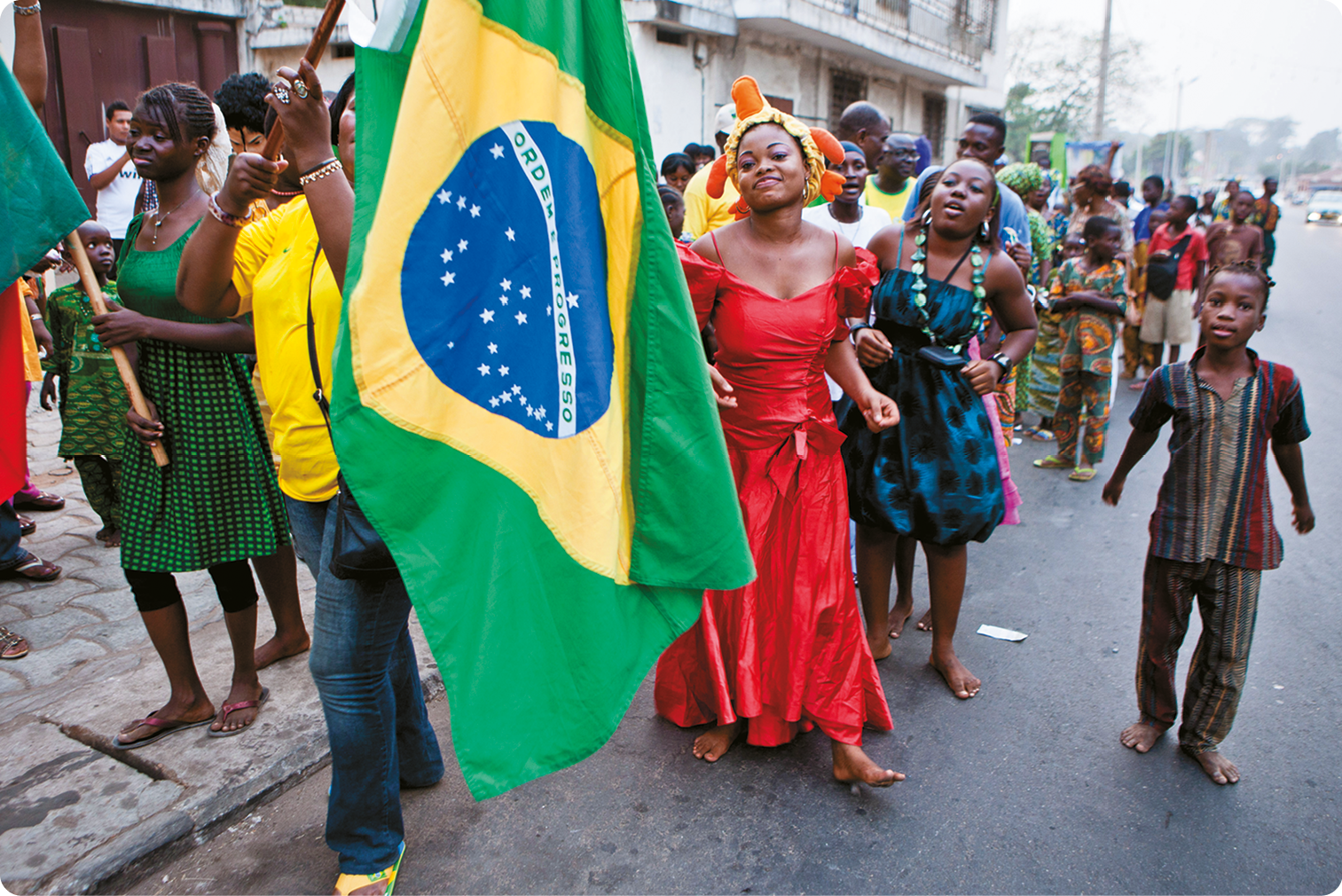 Fotografia. Pessoas negras com roupas coloridas segurando a bandeira do Brasil em uma rua de uma cidade.