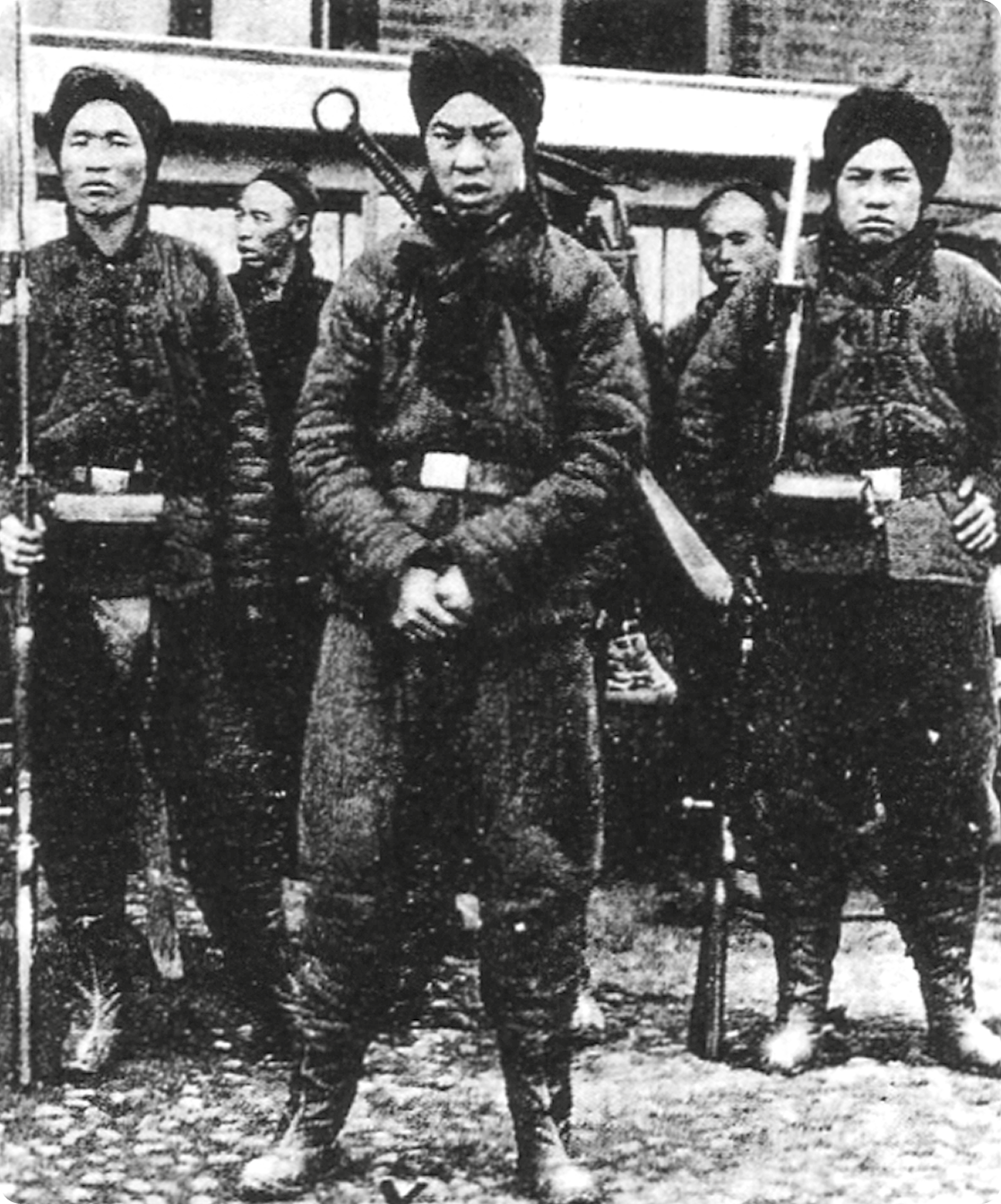 Fotografia em preto e branco. Três homens asiáticos, usando lenço na cabeça e roupas escuras, estão em pé segurando com as mãos bastão.
