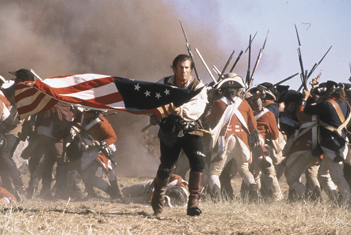 Cena de filme. Um homem correndo e  segurando com as mãos a bandeira dos Estados Unidos. Atrás, soldados com fardas vermelhas, segurando armas com as mãos. No fundo, fumaça.