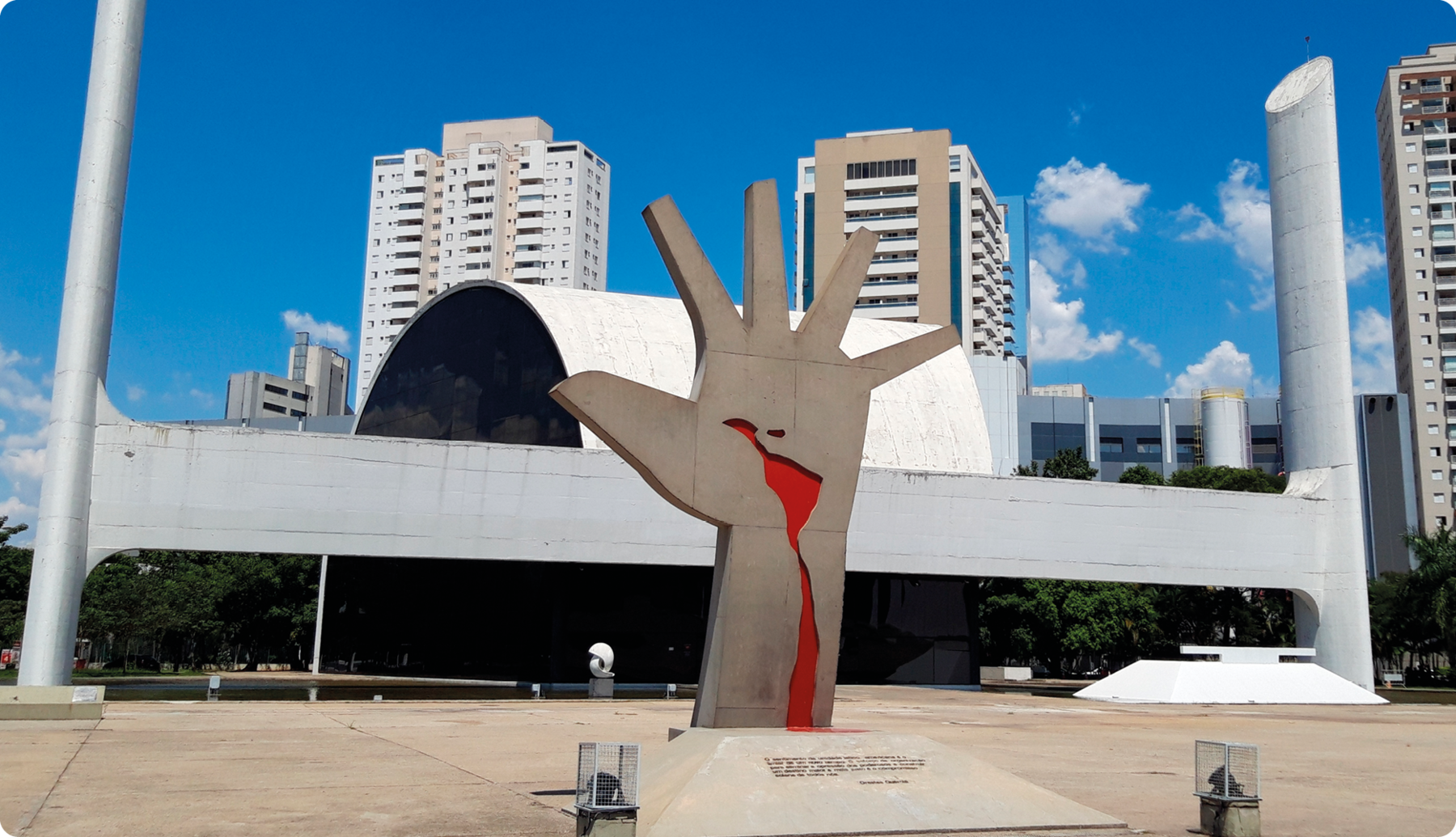 Fotografia. Monumento no formato de uma mão aberta. Na palma da mão, há a silhueta da América Latina e uma linha vermelha que desce até o punho. Atrás, construção com duas colunas e um arco no centro.