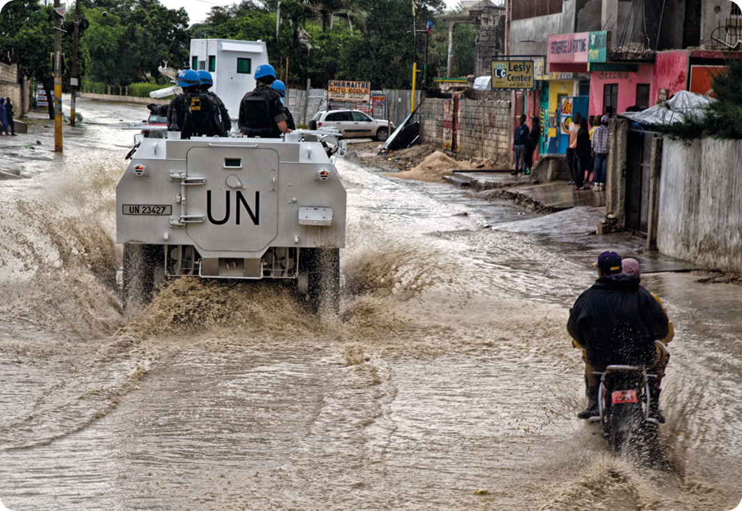 Fotografia. À esquerda, militares em um carro cinza com a sigla UN (United Nations) sobre uma rua alagada. Ao lado, duas pessoas sobre uma moto. Ao redor, moradias.