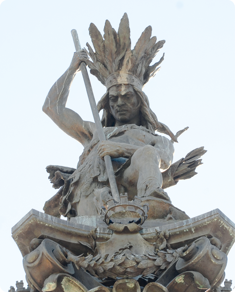 Escultura. Um homem indígena com adorno com plumas na cabeça segurando com as mãos uma lança.