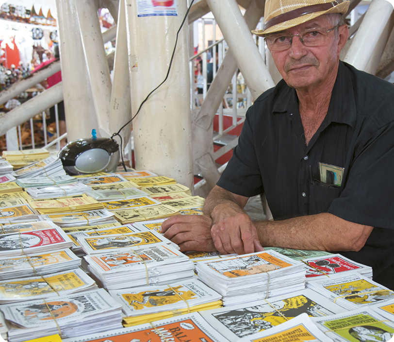 Fotografia. Um homem usando óculos, chapéu e camisa preta, está sentado com as mãos sobre uma mesa onde estão vários livros pequenos expostos em uma banca..