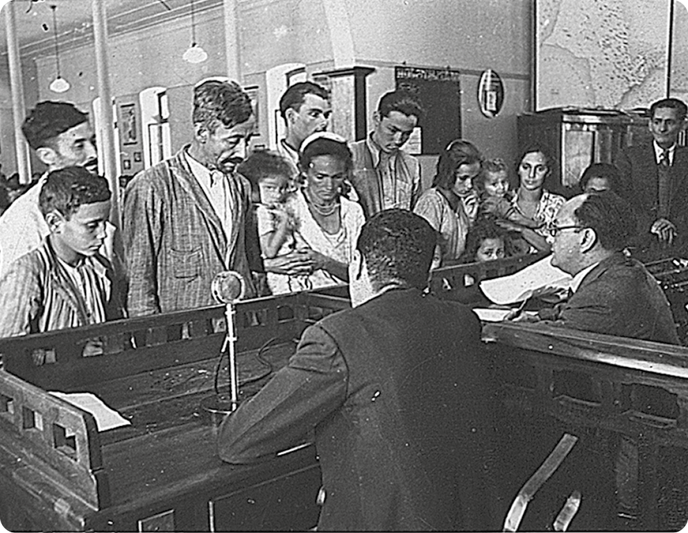 Fotografia em preto e branco. No primeiro plano, um homem de terno está de costas em um balcão. Atrás,  diversas pessoas em pé.