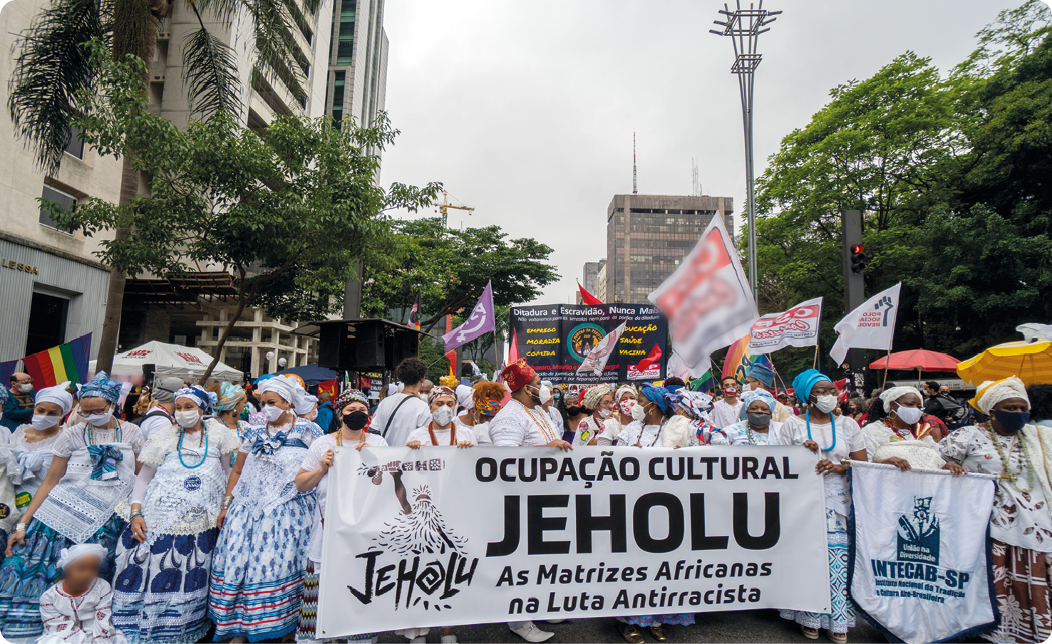 Fotografia. Mulheres negras usando turbante, vestidos rodados, carregando bandeiras e cartazes em uma rua de uma cidade. Ao redor, prédios altos. Em um dos cartazes, há o texto: OCUPAÇÃO CULTURAL JEHOLU. AS MATRIZES AFRICANAS NA LUTA ANTIRRACISTA.