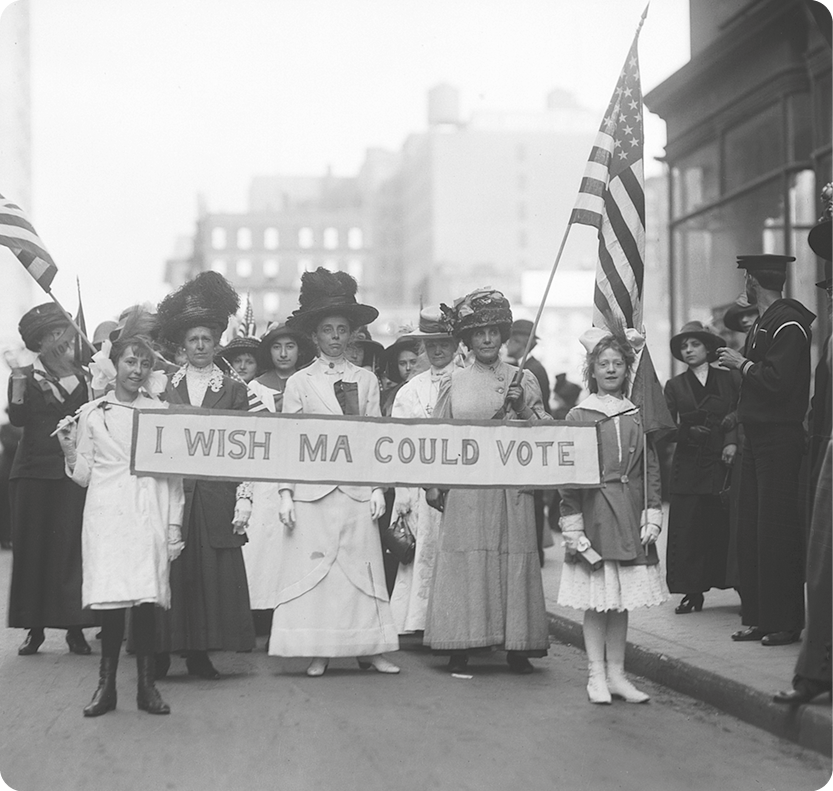 Fotografia em preto e branco. Mulheres e meninas andando em uma rua, segurando bandeira dos Estados Unidos e uma faixa com o texto: I WISH MA COLD VOTE.