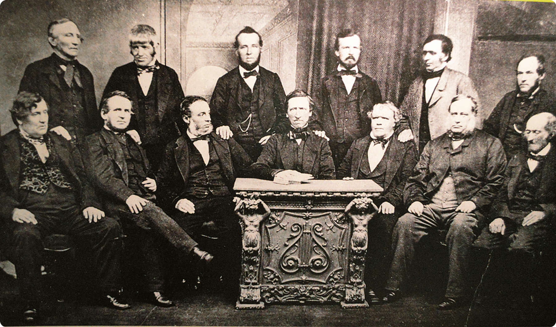 Fotografia em preto e branco. Vários homens usando terno. Alguns estão de pé e outros sentados