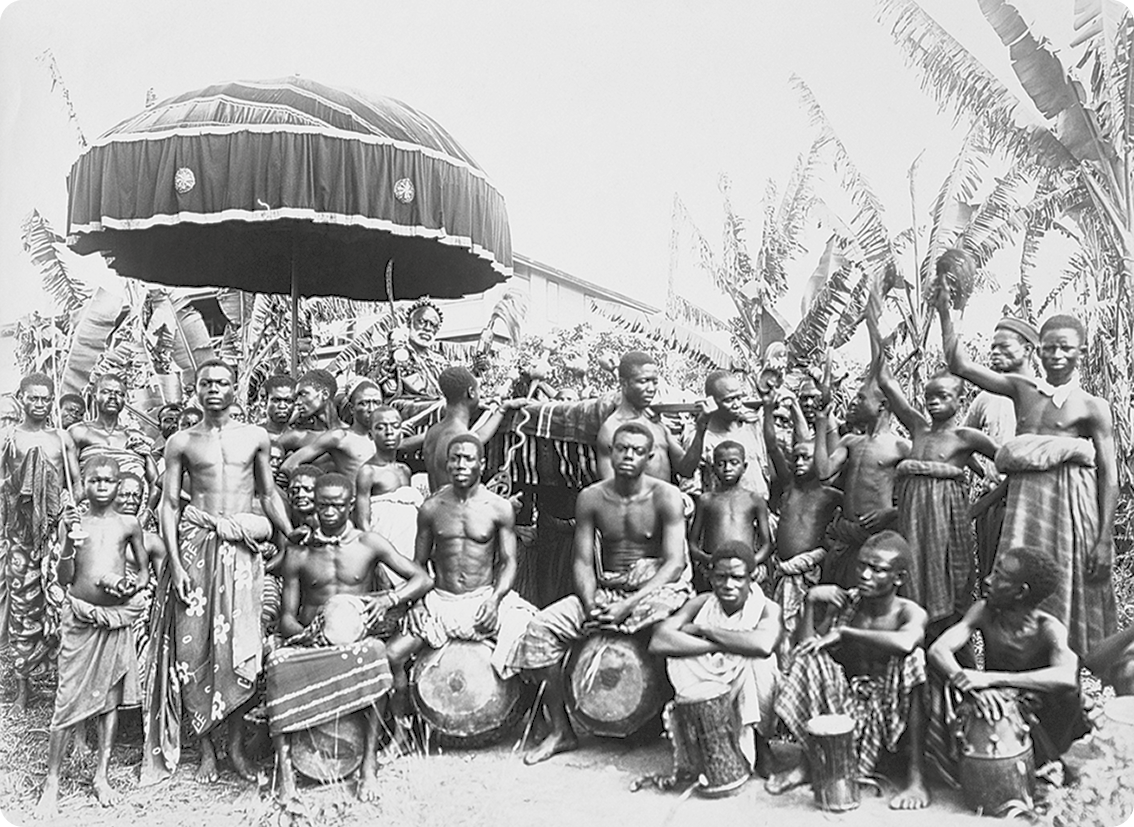 Fotografia em preto e branco. Diversos homens e meninos negros com tecido ao redor da cintura. Alguns estão sentados segurando instrumentos de percussão e outros estão de pé com os braços erguidos. No fundo, vegetação.