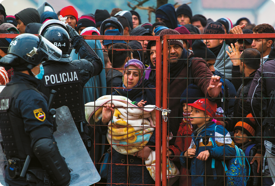 Fotografia. Na frente, dois homens usando farda policiais e capacetes. Eles estão na frente de um portão fechado. Atrás, diversas pessoas, mulheres e crianças.
