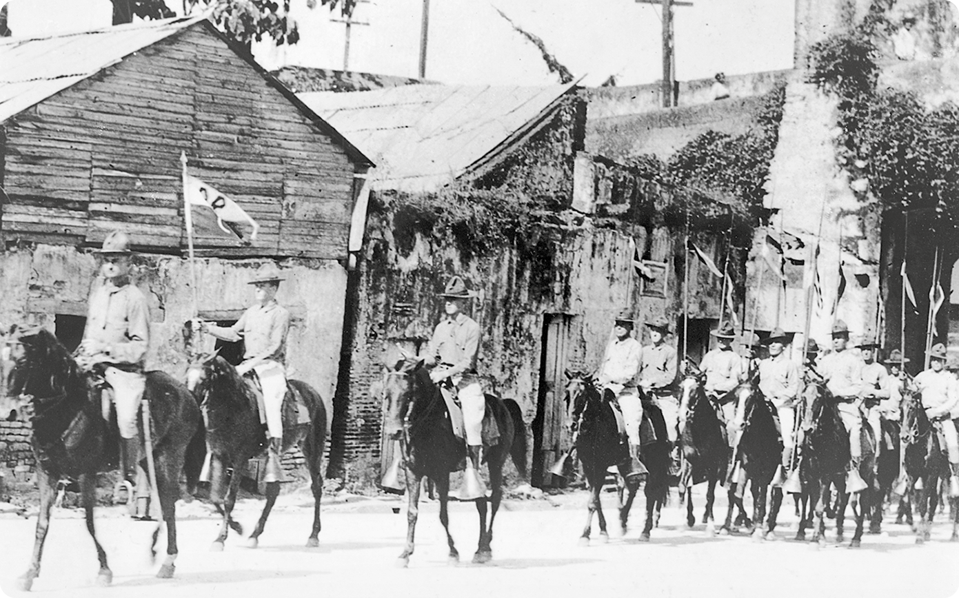 Fotografia em preto e branco. Homens usando chapéu, camisa e calça. Eles estão montados em cavalos, caminhando em uma rua. Atrás, casas de madeiras e de palha.