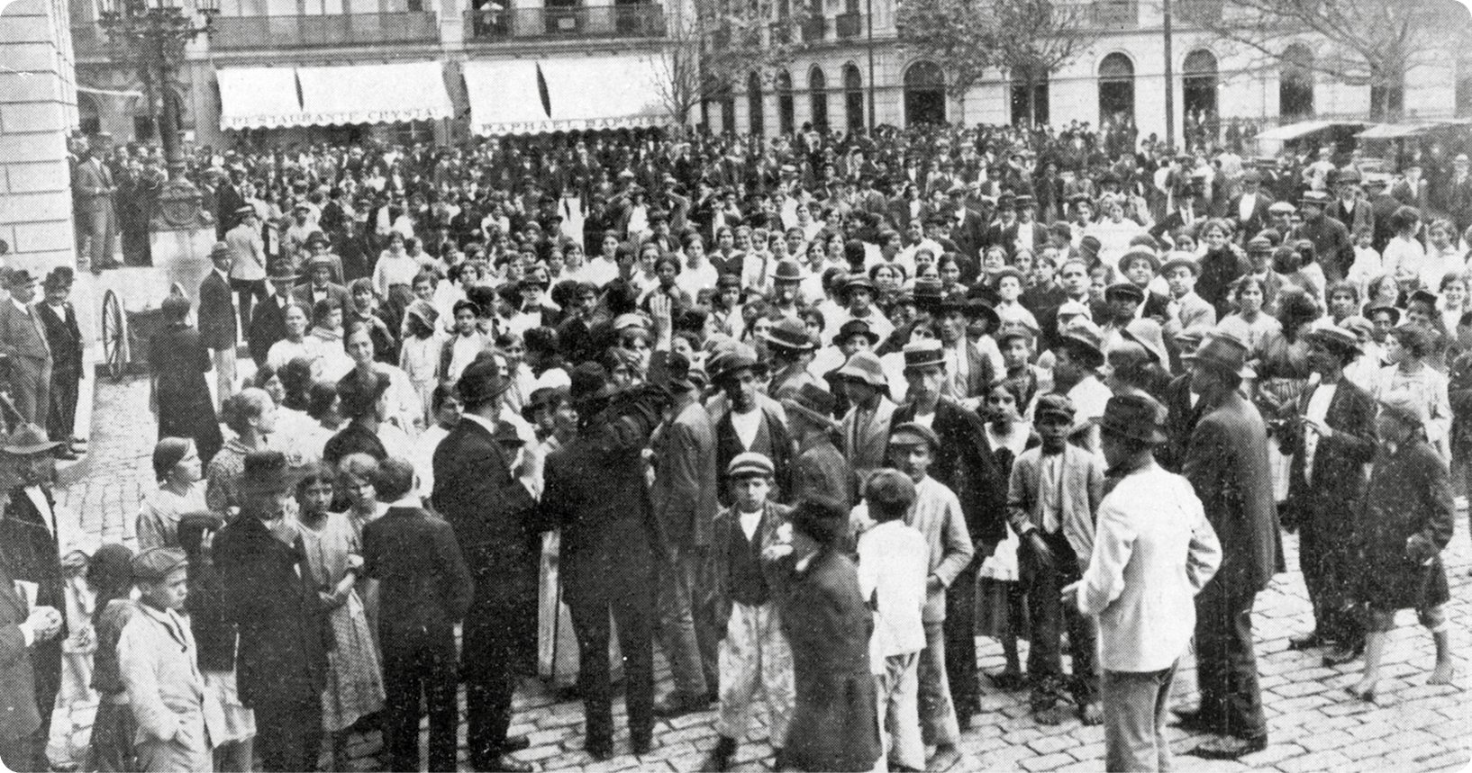 Fotografia em preto e branco. Vista de uma praça. Diversas pessoas, usando chapéu, casaco e vestido, estão em pé em uma rua. À esquerda, pessoas sobre uma estrutura elevada.
