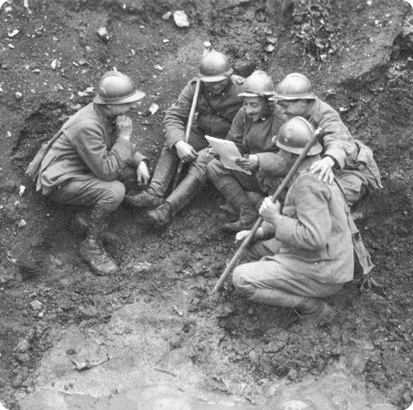 Fotografia em preto e branco. Cinco homens fardados, segurando armas com as mãos. Eles estão sentados na terra, enquanto um deles está segurando uma carta com as mãos.