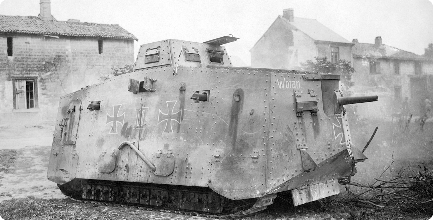 Fotografia em preto e branco. Um tanque de guerra, com ilustração de duas cruzes na lateral.  Atrás, algumas moradias em ruínas.