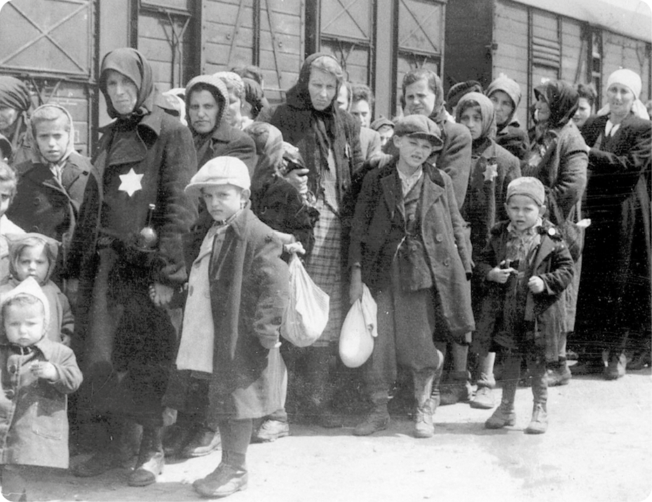 Fotografia em preto e branco. Mulheres e crianças usando a estrela de Davi no peito. Elas estão em pé, em fila e ao lado de um trem.