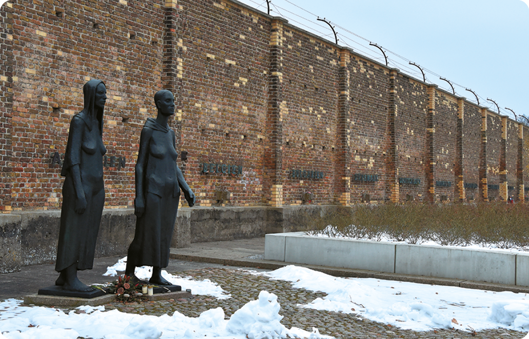 Escultura. Duas mulheres em pé. Atrás uma muralha de tijolos.
