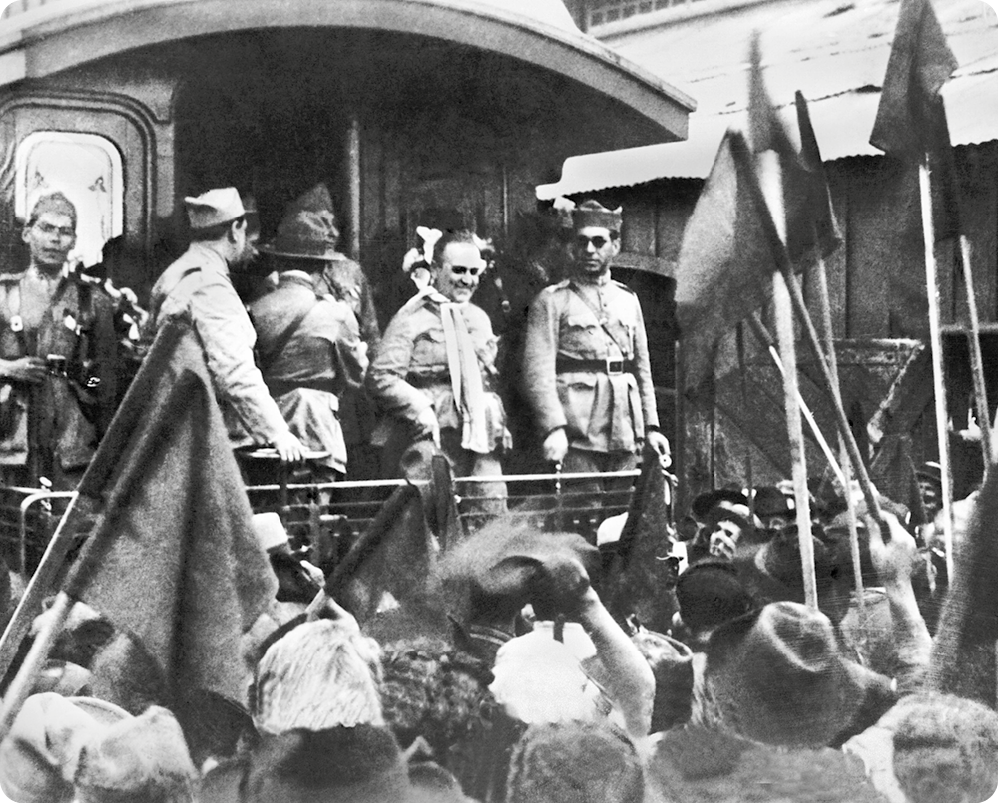 Fotografia em preto e branco. Ao centro, em cima de um trem, homens usando roupas militares. Ao redor, varias pessoas estão em pé, olhando os homens fardados no trem. Algumas delas seguram bandeiras