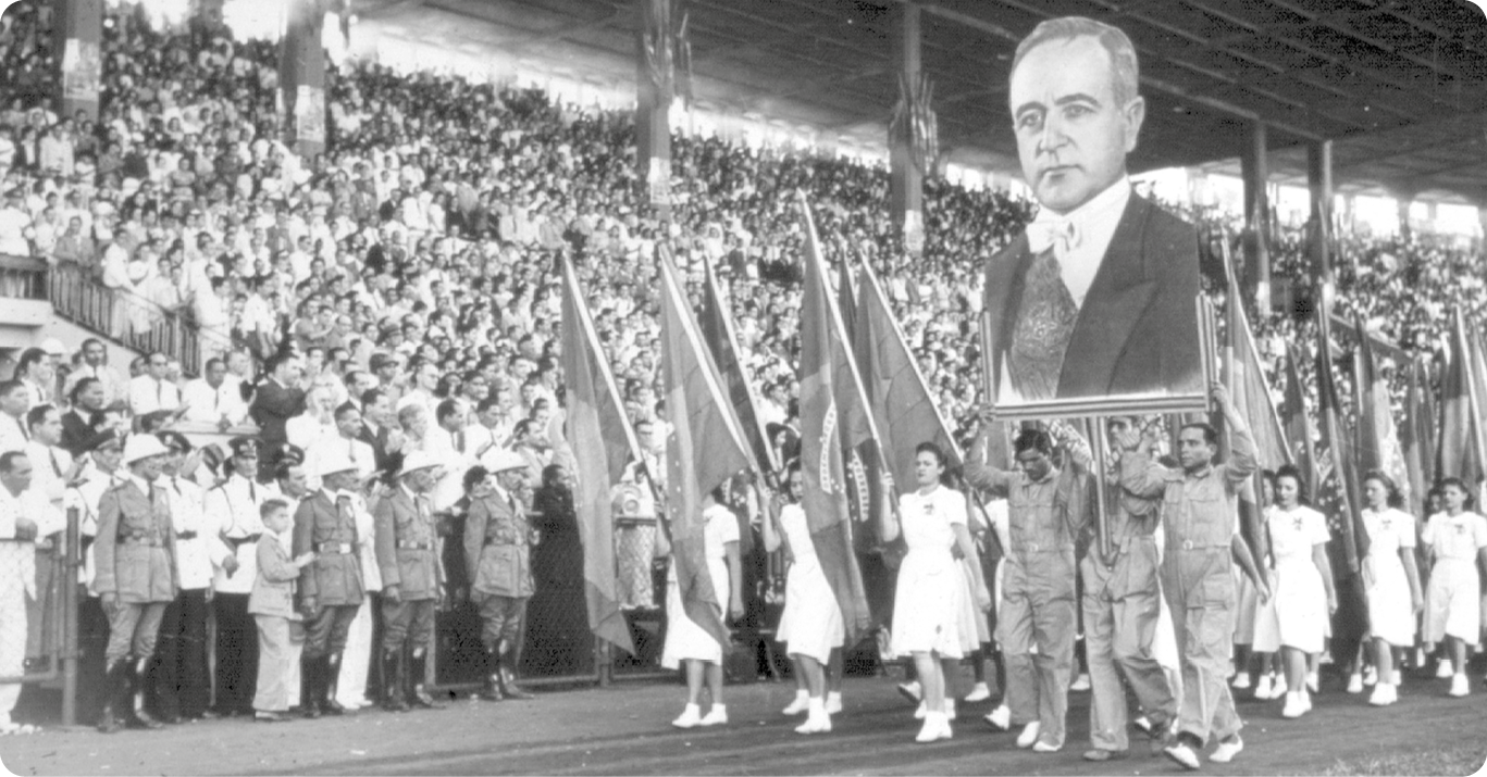 Fotografia em preto e branco. No primeiro plano, mulheres usando vestidos e segurando bandeiras do Brasil. Ao centro, três homens segurando um cartaz com o busto de um homem usando terno e uma faixa. Atrás, diversas pessoas em pé sobre uma arquibancada.