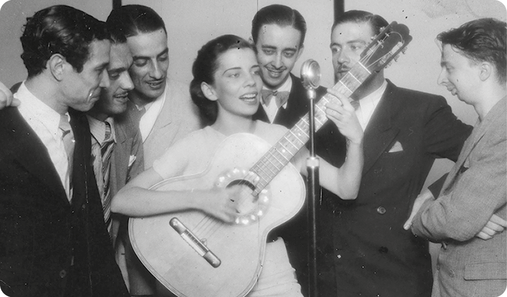 Fotografia em preto e branco. Ao centro, mulher, com cabelos presos e usando vestido, está segurando com as mãos um violão e com a boca próxima a um microfone. Ao redor, homens usando terno.