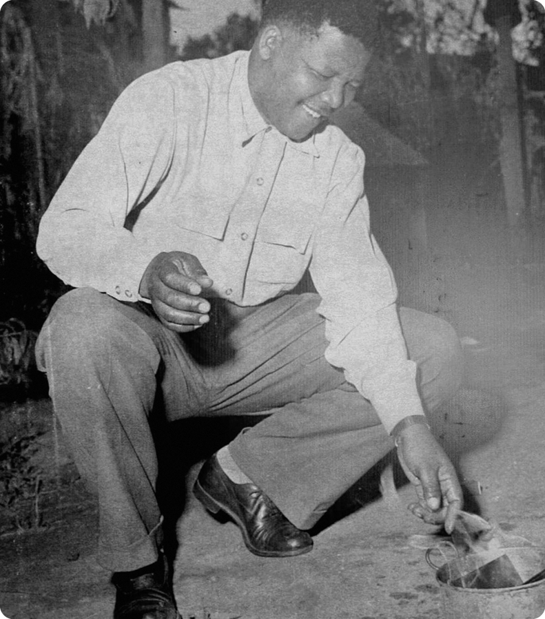 Fotografia em preto e branco. Um homem negro usando camisa com botões e calça. Ele está agachado com as mãos perto de uma fogueira no chão. Ele sorri.