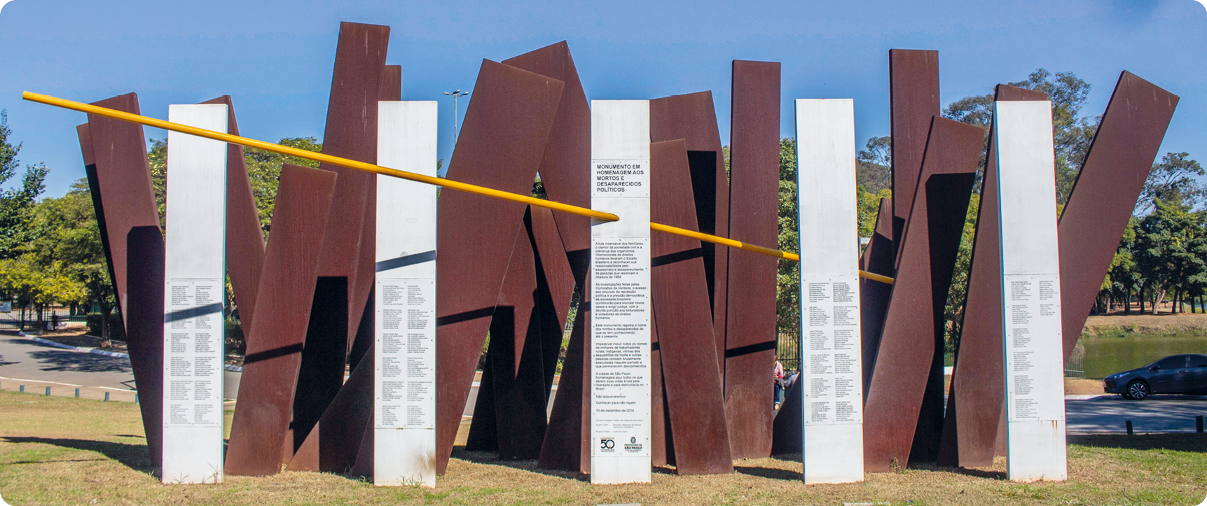 Fotografia. Monumento composto por colunas de metal cruzadas com textos sobre elas. Uma haste amarela atravessa as colunas.