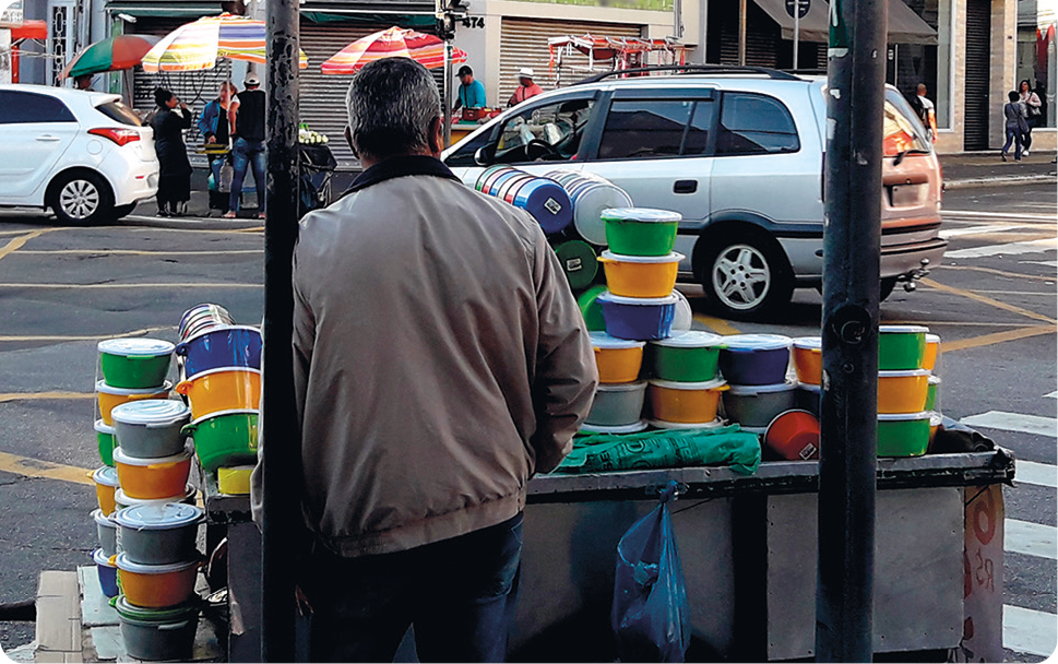 Fotografia. No primeiro plano, um homem, usando casaco, está de costas e encostado em uma barraca com vários potes coloridos. Ao fundo, rua com vários carros..