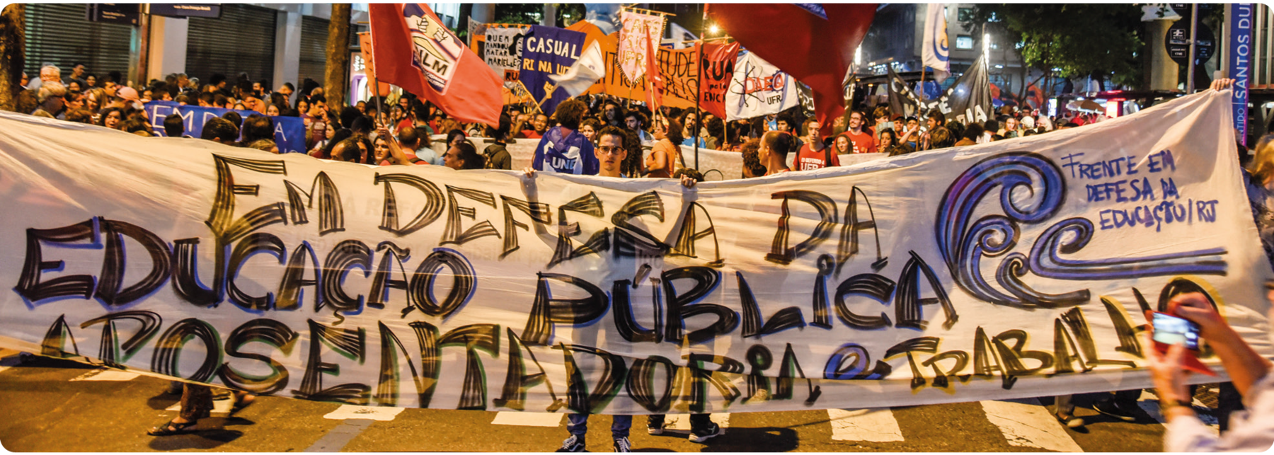 Fotografia. Pessoas segurando placas e cartazes em uma rua. Ao centro, o cartaz com o seguinte texto: EM DEFESA DA EDUCAÇÃO PÚBLICA. APOSENTADORIA E TRABALHO. FRENTE EM DEFESA DA EDUCAÇÃO. RIO DE JANEIRO.