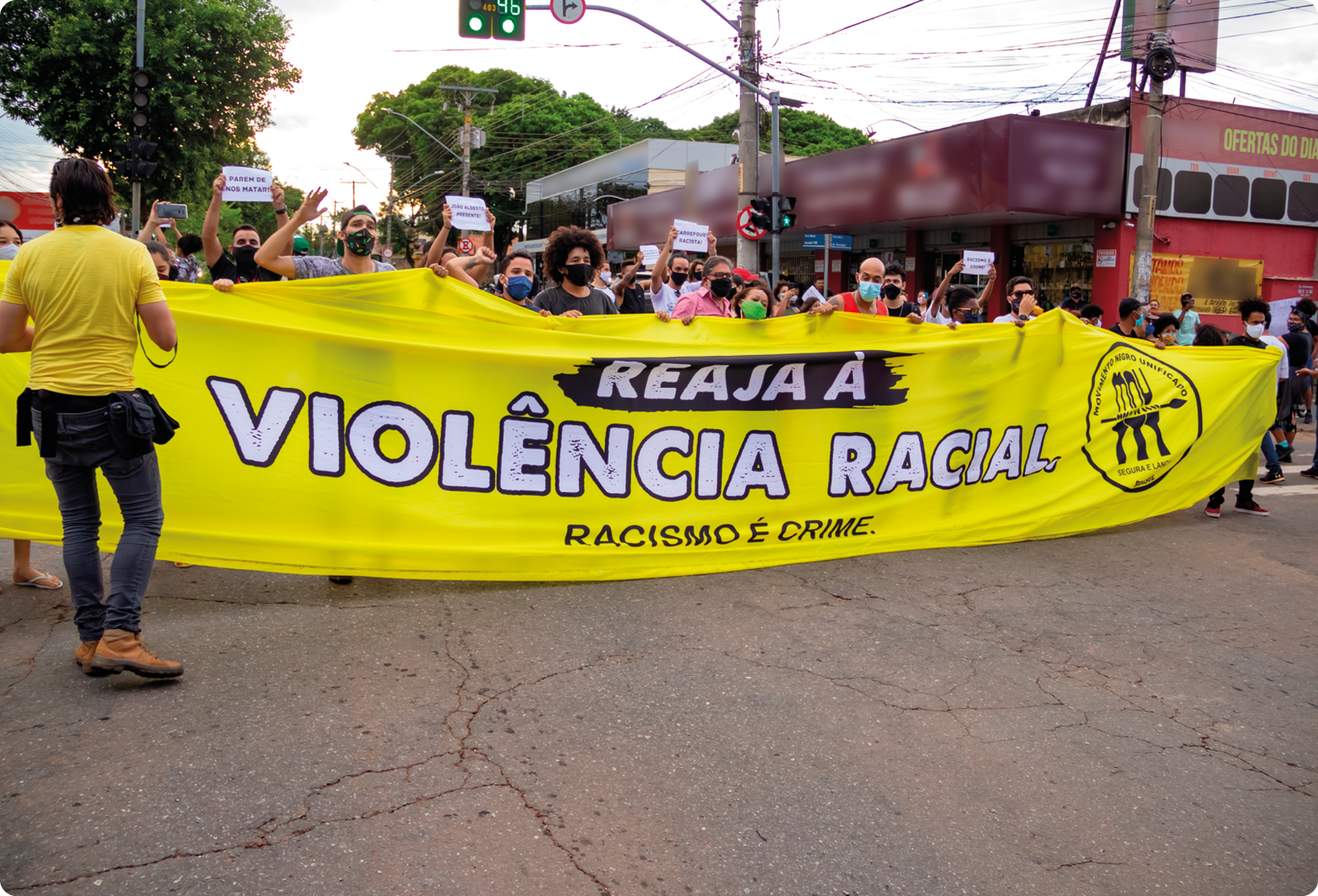 Fotografia. Pessoas em uma rua segurando uma faixa com o texto: REAJA À VIOLÊNCIA RACIAL. RACISMO É CRIME.