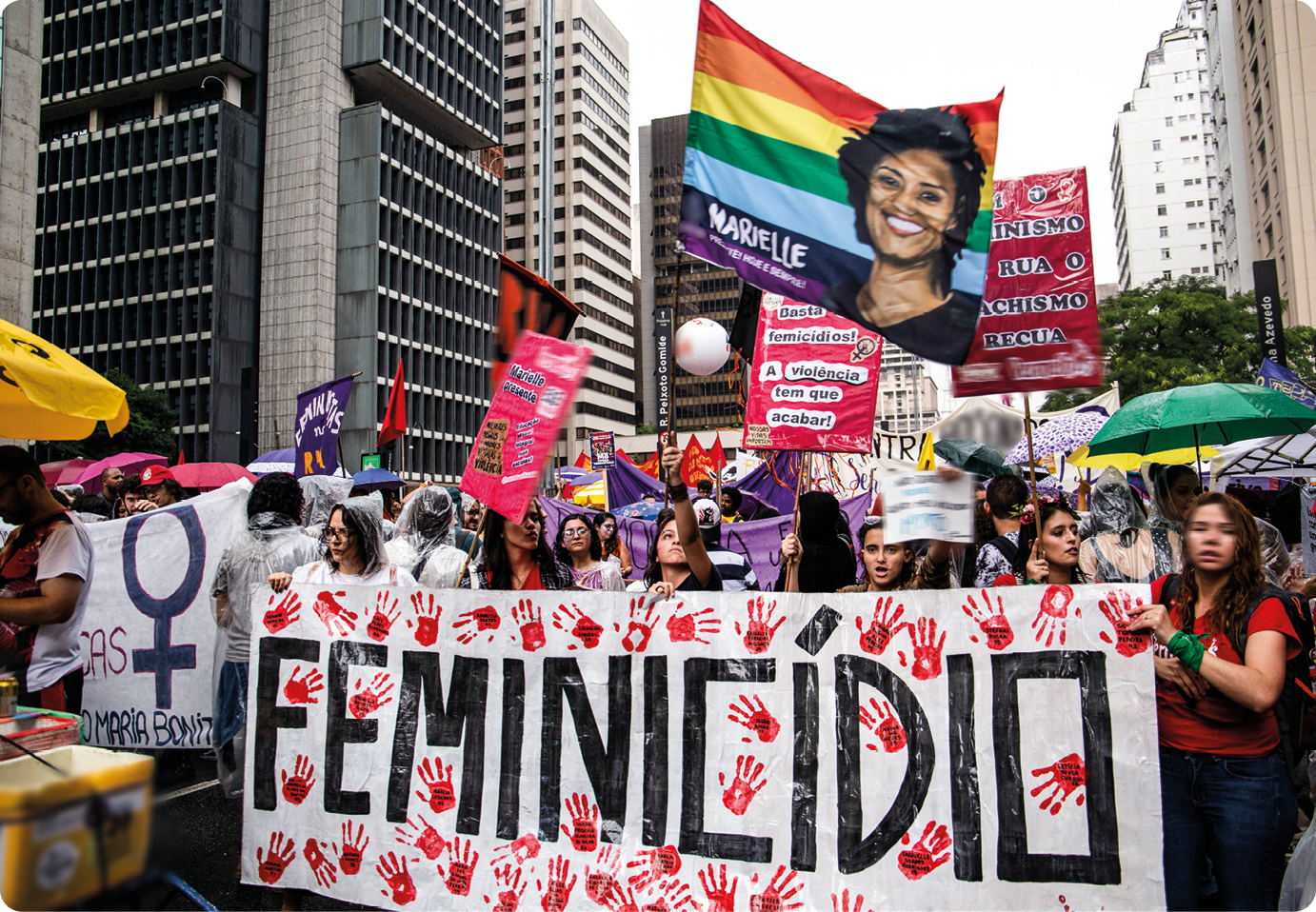Fotografia. Diversas mulheres em uma rua, segurando bandeiras, faixas e cartazes. Ao centro, cartaz com o texto: FEMINICÍDIO.