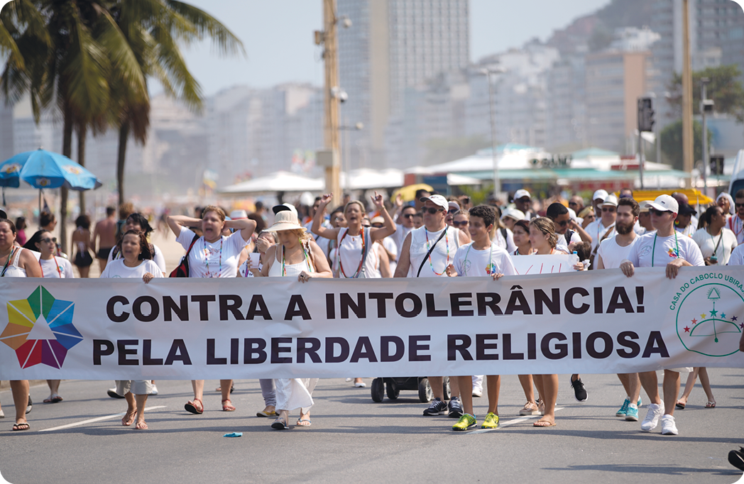 Fotografia. Diversa pessoas, com roupas brancas, caminhando em uma rua e segurando um cartaz com o texto: CONTRA A INTOLERÂNCIA! PELA LIBERDADE RELIGIOSA.