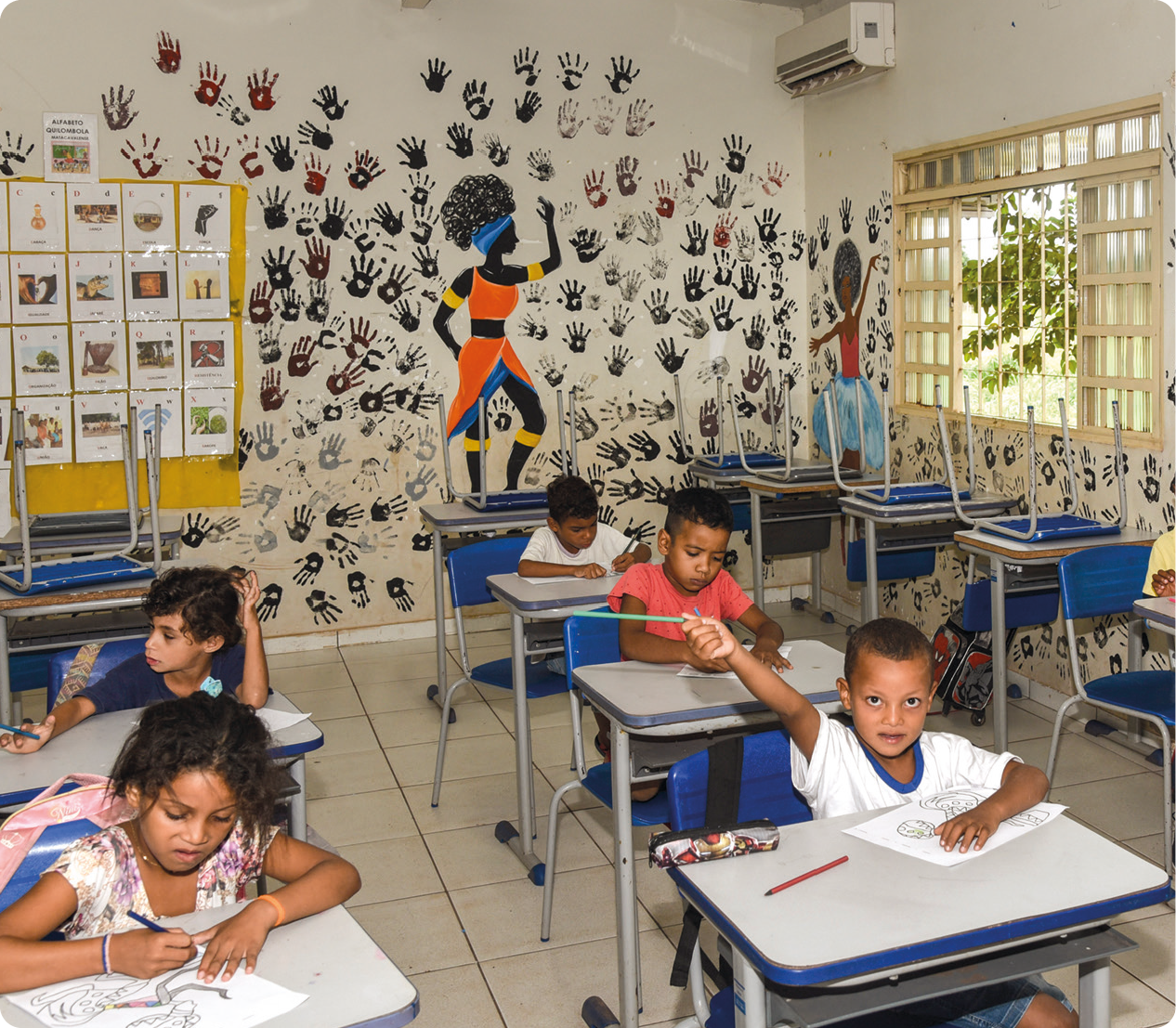 Fotografia. Vista de uma sala de aula. No primeiro plano, algumas crianças sentadas e pintando desenhos. Ao fundo, parede com ilustração de mulheres negras e silhuetas de mãos