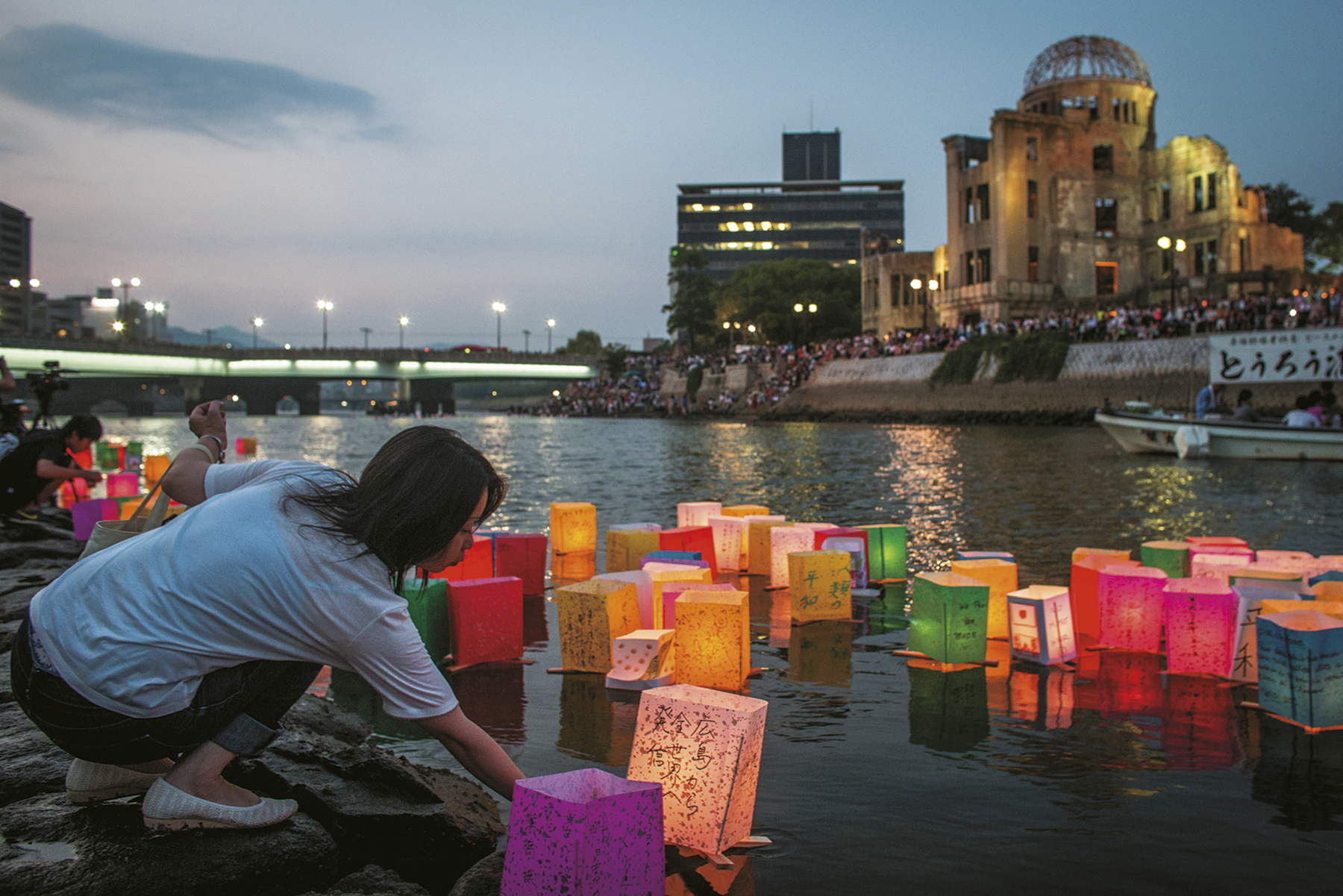 Fotografia. Uma mulher asiática, usando camisa e calça, está agachada as margens de um rio, onde há diversas lanternas com texto escrito no idioma japonês. No fundo, ruína de um edifício.