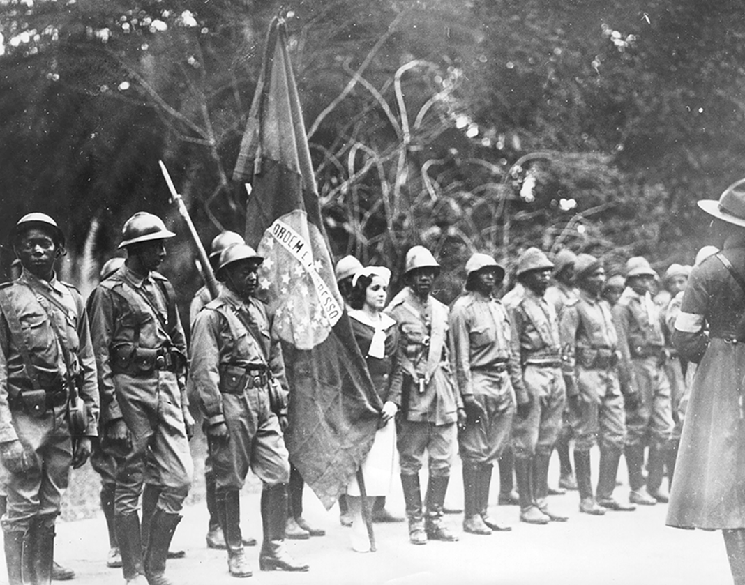 Fotografia em preto e branco. Soldados negros, um do lado do outro, usando fardas militares. Alguns deles estão segurando armas. Ao centro, uma mulher branca está de pé e segurando a bandeira do Brasil.