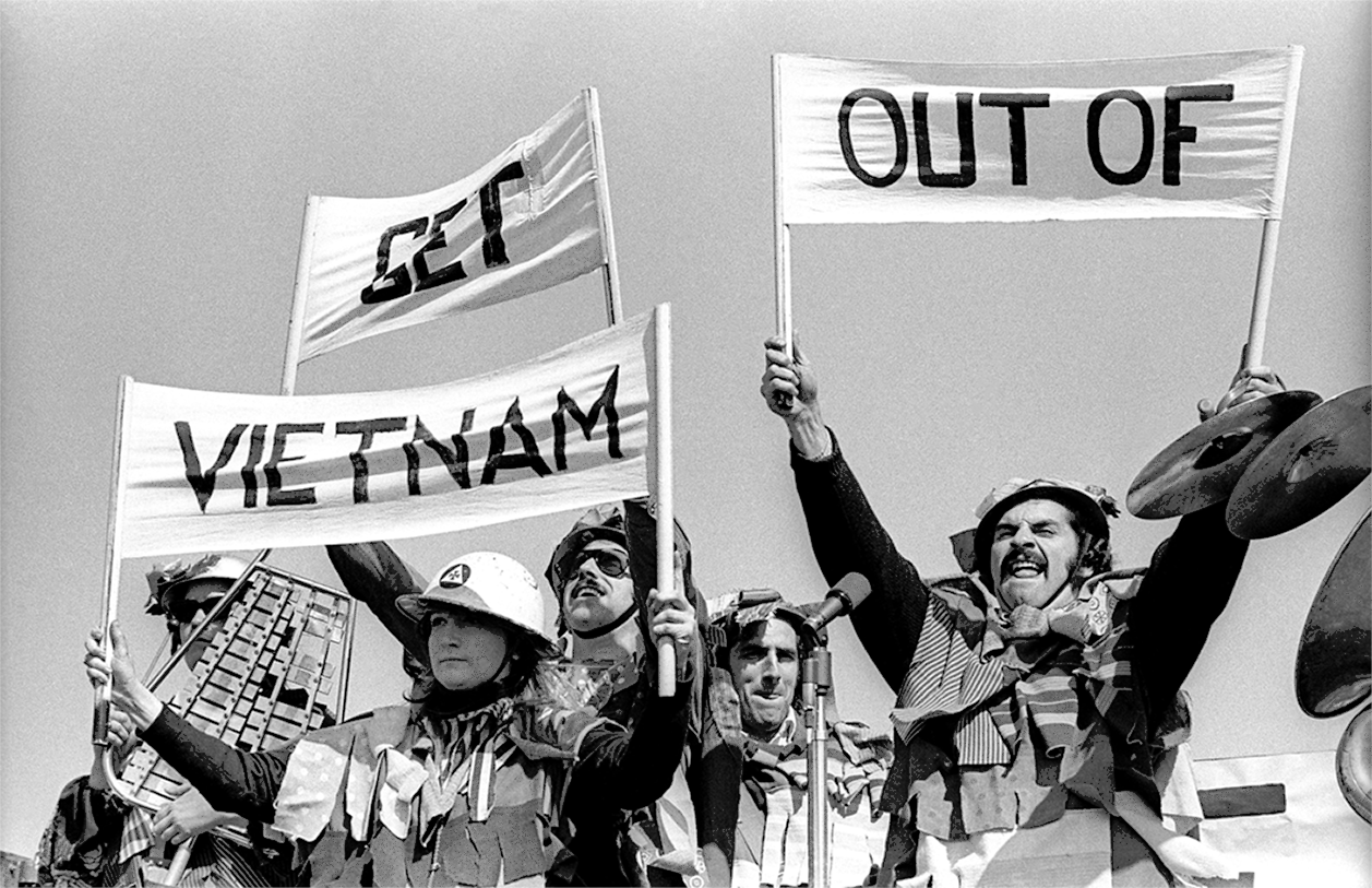 Fotografia em preto e branco. Pessoas, fantasiadas com camuflagem, capacetes e outros objetos, estão segurando cartazes com o texto: GET VIETNAM OUT OF.