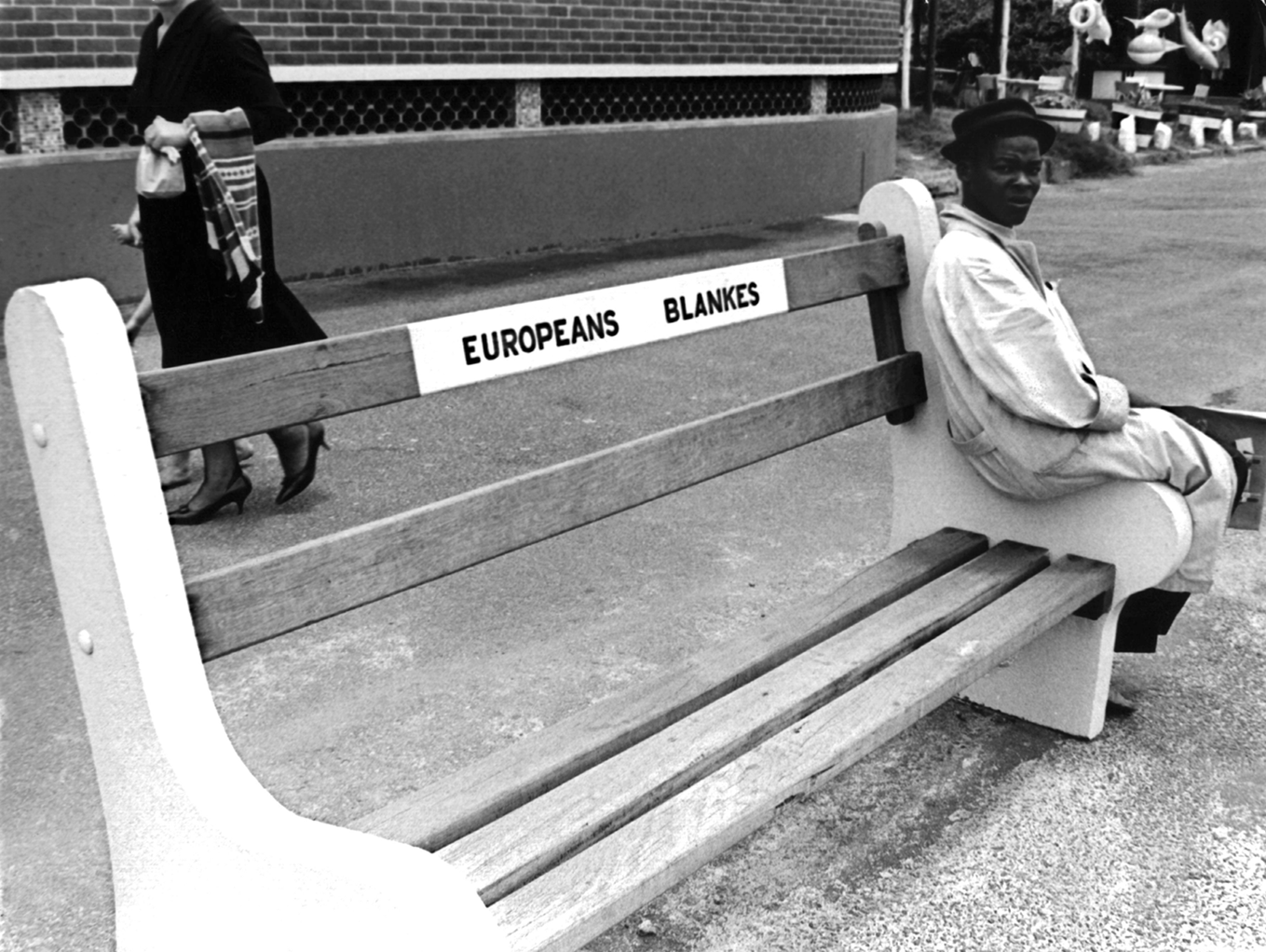 Fotografia em preto e branco. Destacando um banco em uma calçada. No cento do banco, o texto: EUROPEANS BLANKES. À direita, um homem negro sentado na ponta do banco.