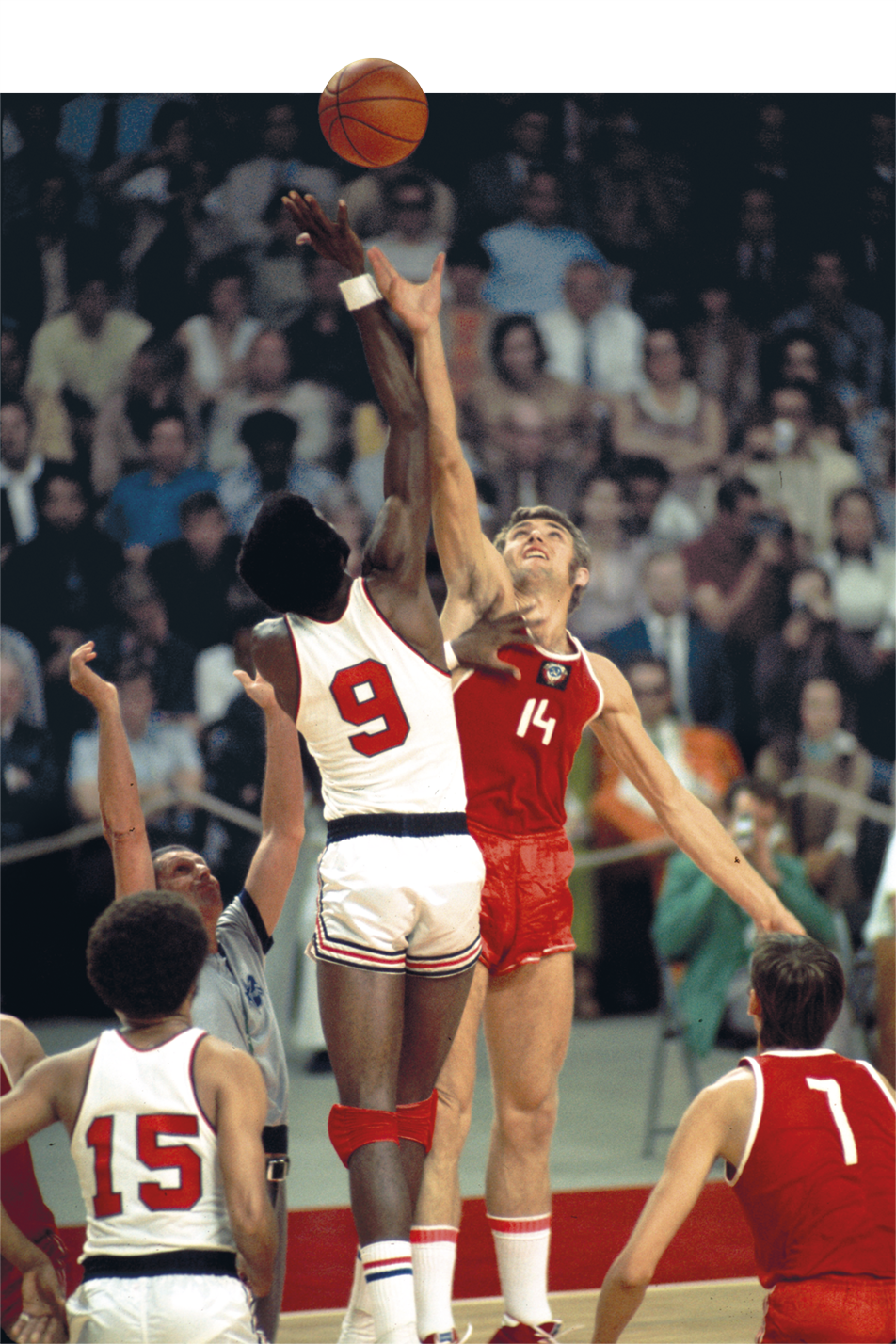 Fotografia. Dois homens, um usando uniforme de basquete branco e o outro com uniforme vermelho, estão com um dos braços estendidos na direção de uma bola. Ao fundo, pessoas assistindo.