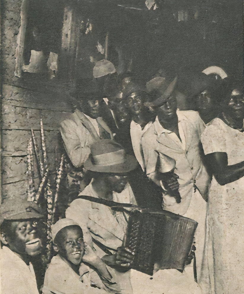 Fotografia em preto e branco. À esquerda, dois jovens negros, sentados e sorrindo. Ao lado, um homem negro, usando chapéu e roupas brancas, segurando um acordeão. À direita, homens e mulheres negras batendo palmas.