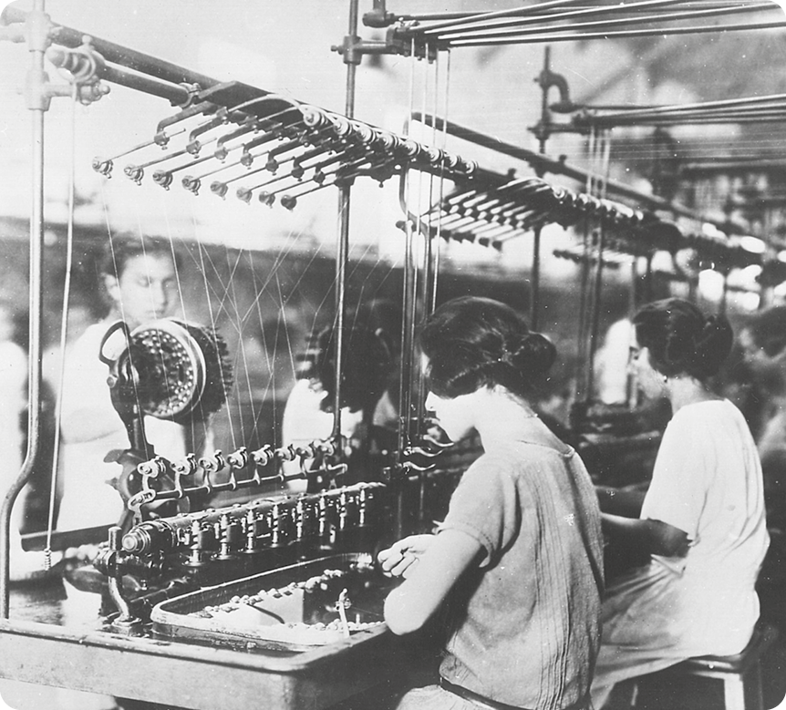 Fotografia em preto e branco. Duas mulheres com cabelos na altura da orelha, usando vestido. Elas estão sentadas com as mãos sobre uma máquina de tecido. Atrás, um homem usando terno. Ele está de pé.
