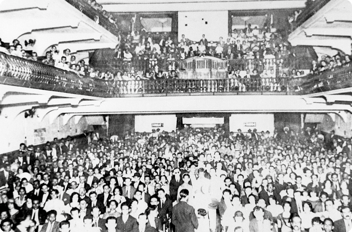 Fotografia em preto e branco. Vista de uma sala grande com mezanino. Há diversas pessoas sentadas.