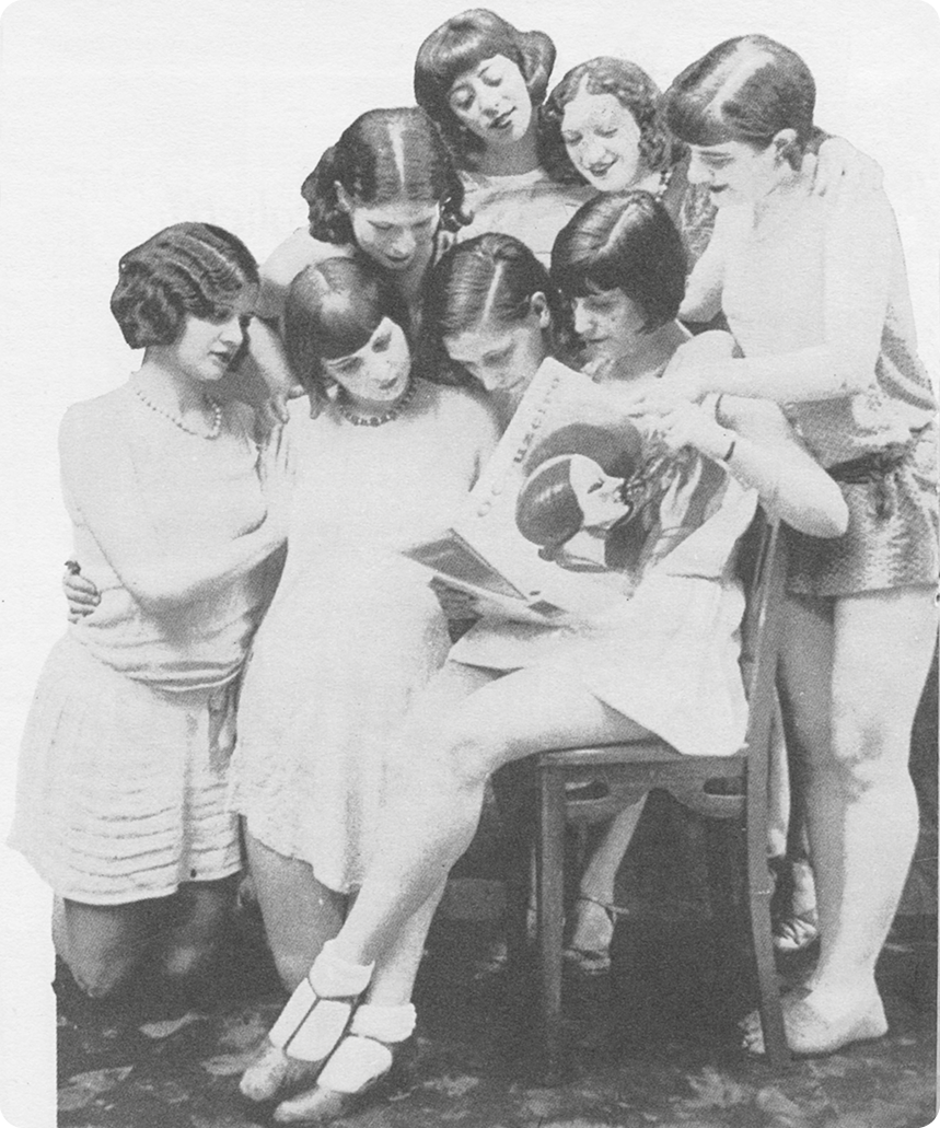 Fotografia em preto e branco. Ao centro, uma mulher com cabelos na altura das orelhas e usando um vestido. Ela está sentada em uma cadeira segurando com as mãos uma revista. Ao redor, sete mulheres jovens olhando na direção da revista.