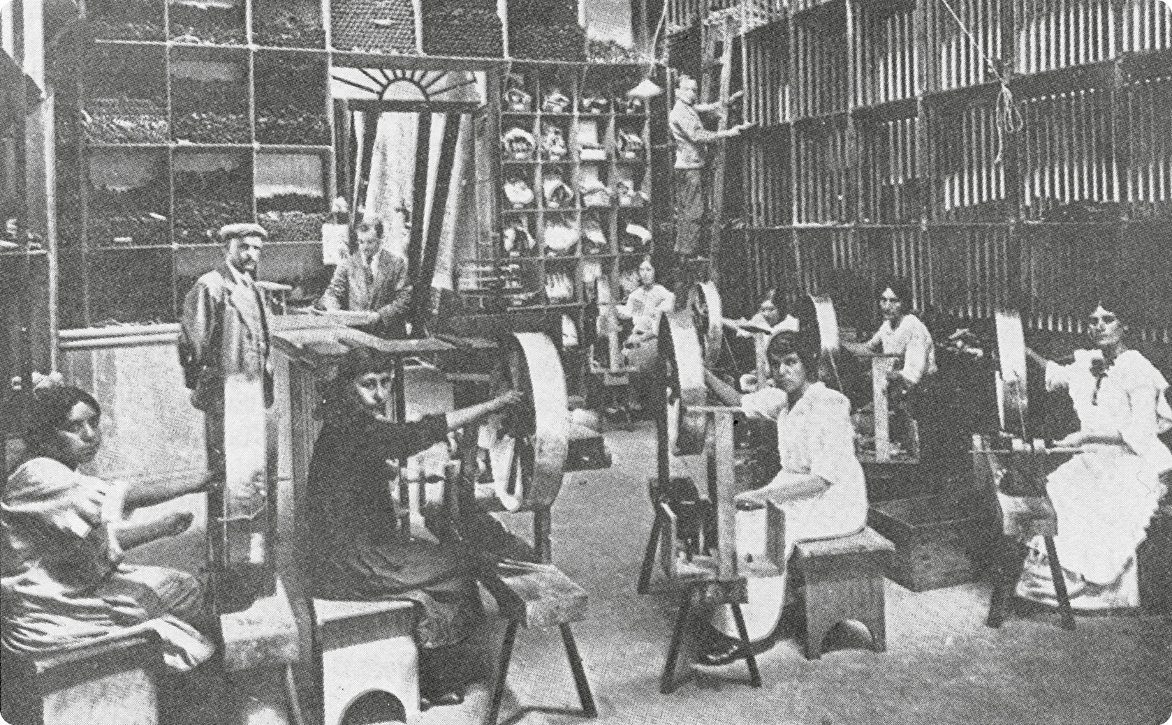 Fotografia em preto e branco. No primeiro plano, mulheres usando vestido, sentadas em bancos e com as mãos sobre uma pequena máquina de tecido. Ao fundo, homens, com terno, em pé.