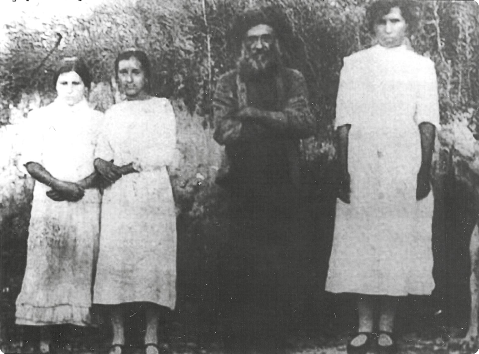 Fotografia em preto e branco. À esquerda, duas mulheres, usando vestidos brancos, estão em pé  com as mãos na frente do corpo. Ao lado, um homem, usando chapéu e túnica escura, está com os braços cruzados. À direita, uma mulher usando vestido.
