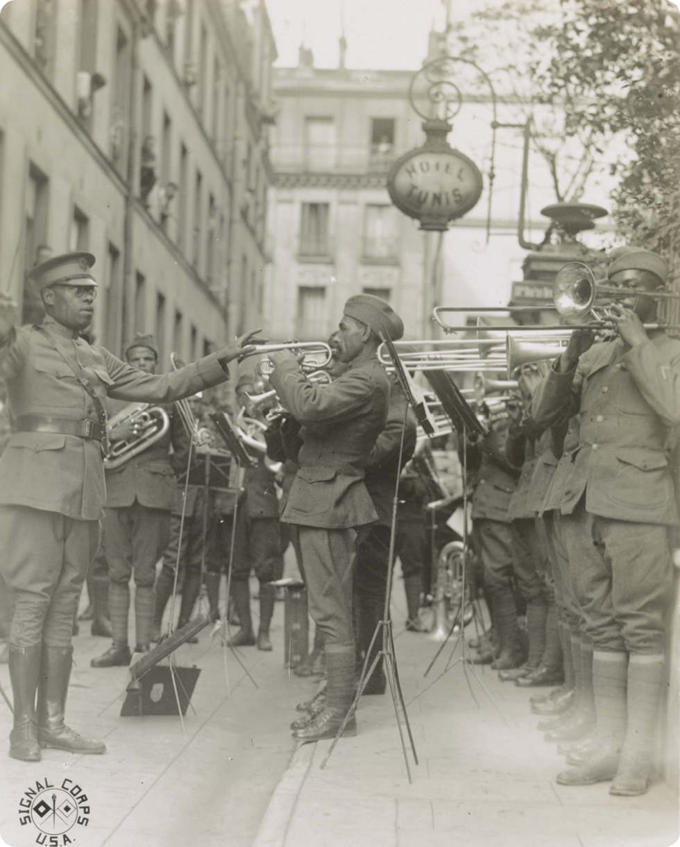 Fotografia em preto e branco. Homens negros, usando chapéu e farda, estão em pé tocando instrumentos de sopro.