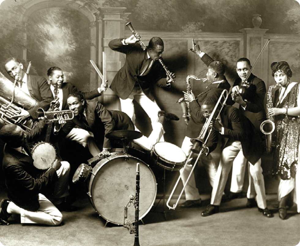 Fotografia em preto e branco. Homens, na sua maioria negros, vestindo terno e gravata, e uma mulher negra, usando vestido, estão tocando instrumentos de sopro e percussão.