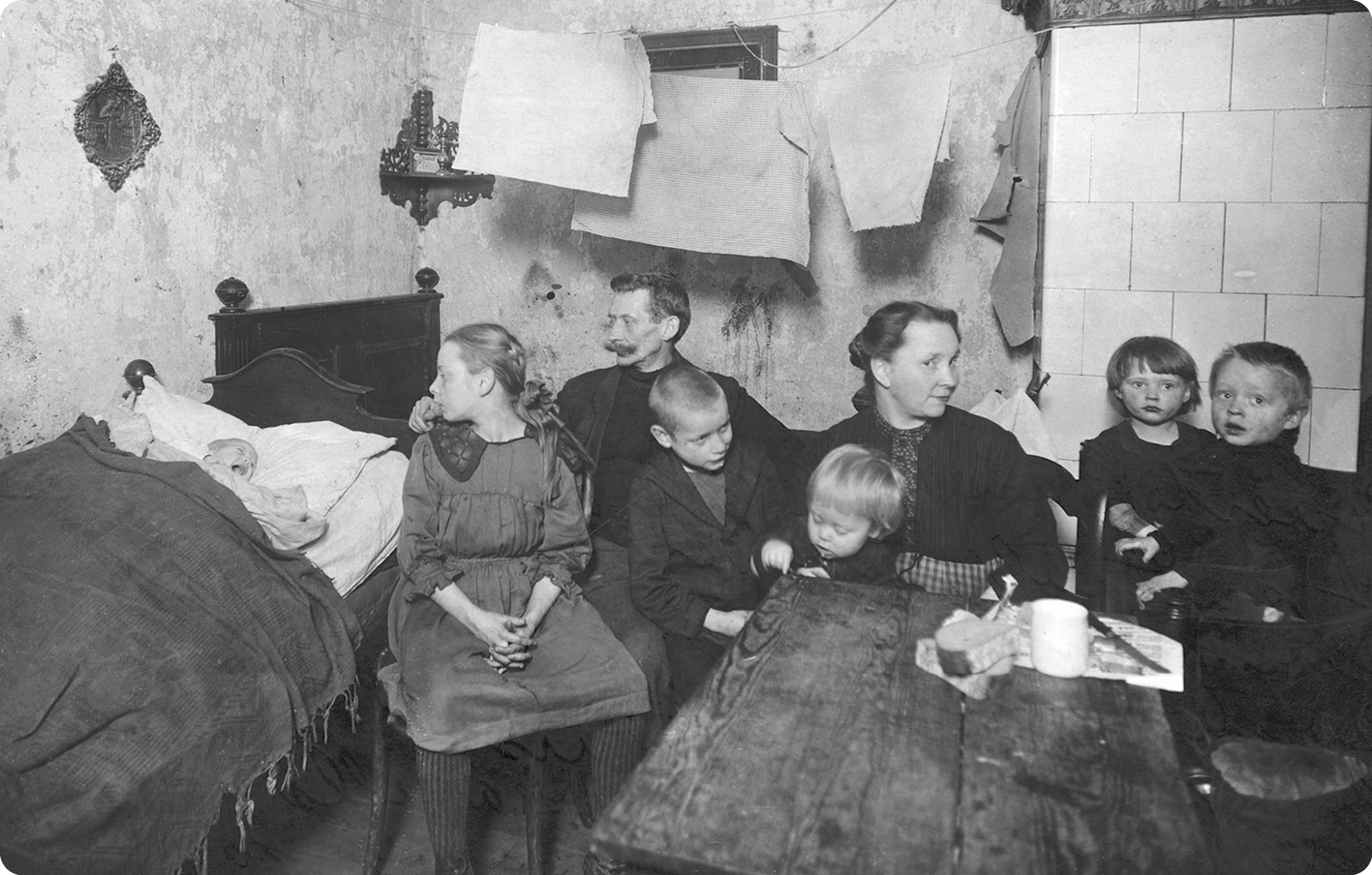 Fotografia em preto e branco. À esquerda uma cama. Sobre a cama, um bebê com a boca aberta. Ao lado, uma menina, um homem, uma mulher e quatro crianças estão sentadas ao redor de uma mesa pequena.