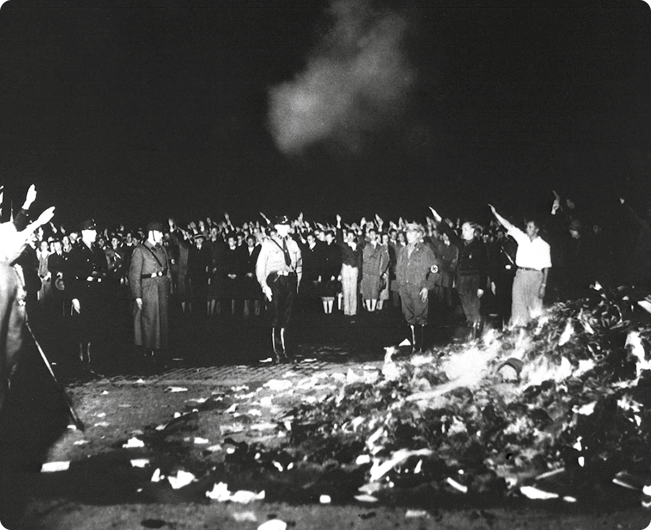 Fotografia em preto e branco. Diversas pessoas em uma rua. Elas estão com uma das mãos levantadas na posição diagonal. Centralizado, uma fogueira com livros.