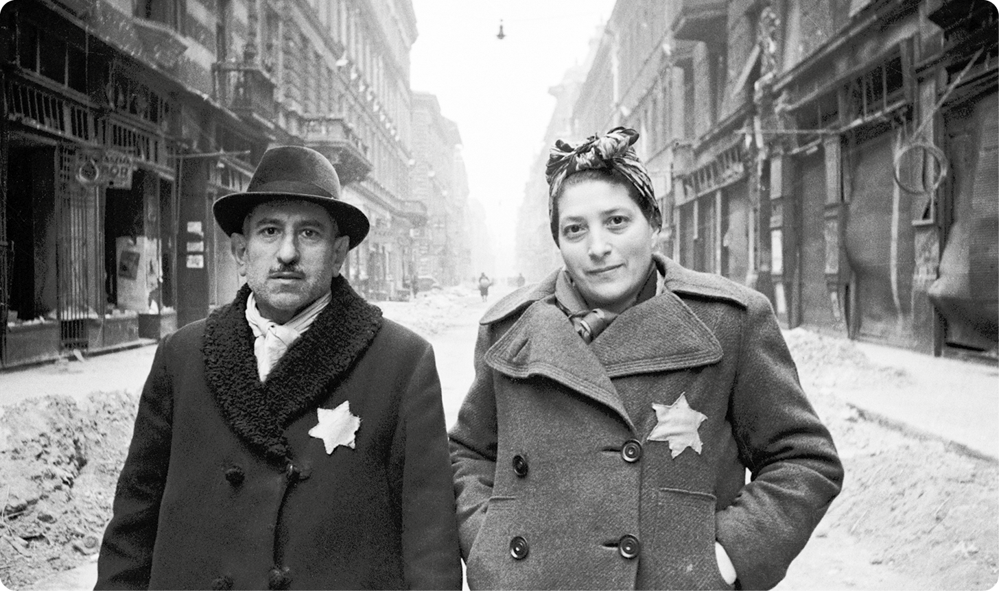 Fotografia em preto e branco. À esquerda, um homem usando chapéu e casaco, com uma estrela de Davi no peito. Ao lado, uma mulher com um lenço na cabeça, usando um sobretudo com uma estrela de Davi. Eles estão de pé em uma rua com neve ao redor.