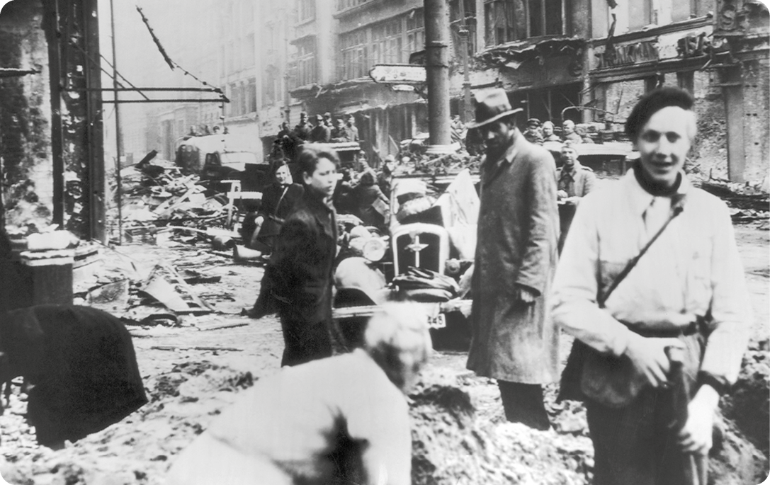 Fotografia em preto e branco. Pessoas caminhando em uma rua com edifícios destruídos.