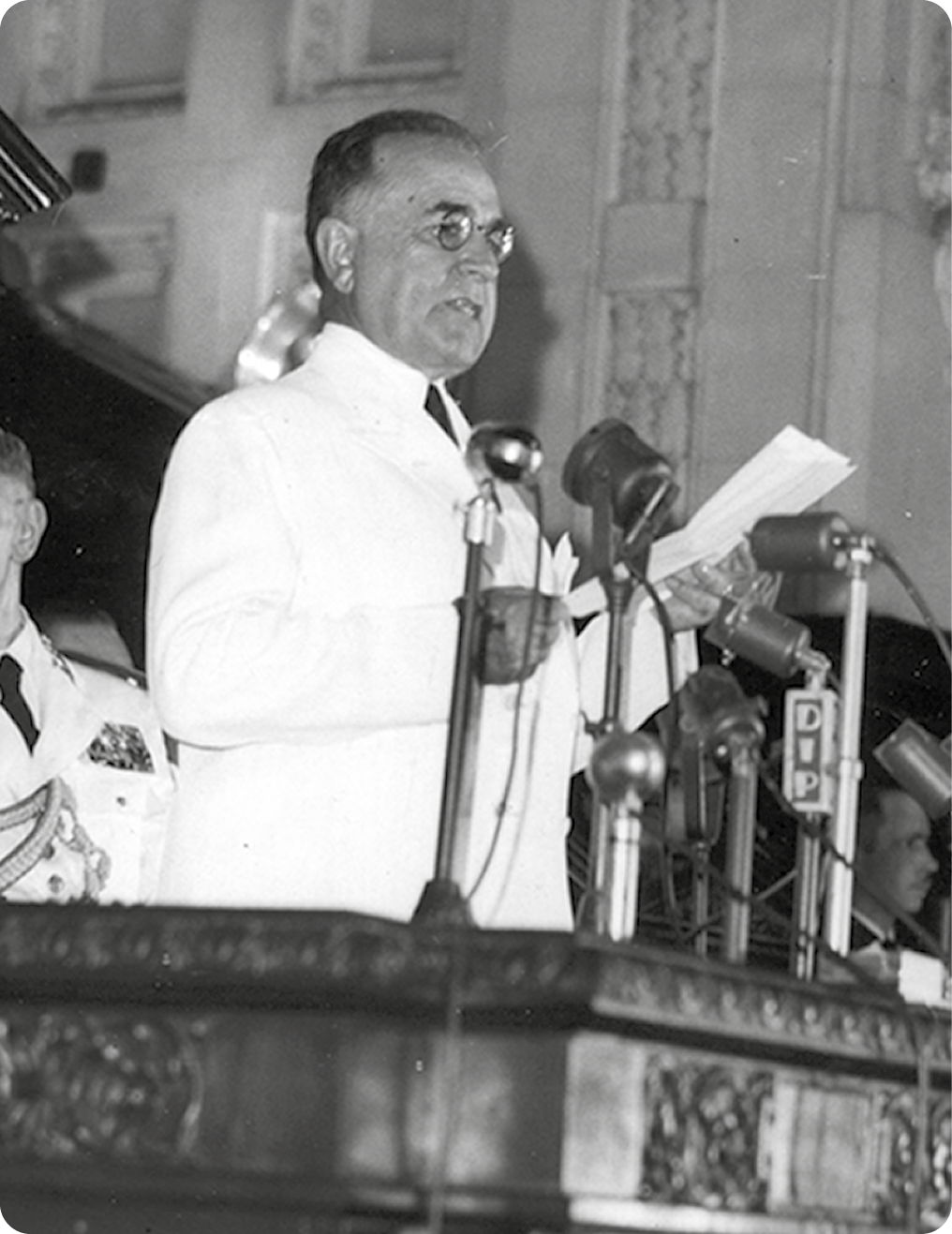 Fotografia em preto e branco. Homem com cabelos curtos, usando óculos e terno branco, está de pé em um púlpito com microfones e segurando uma folha de papel com as mãos.